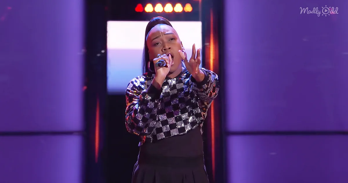 A person in a checkered jacket sings into a microphone on stage with purple lighting.