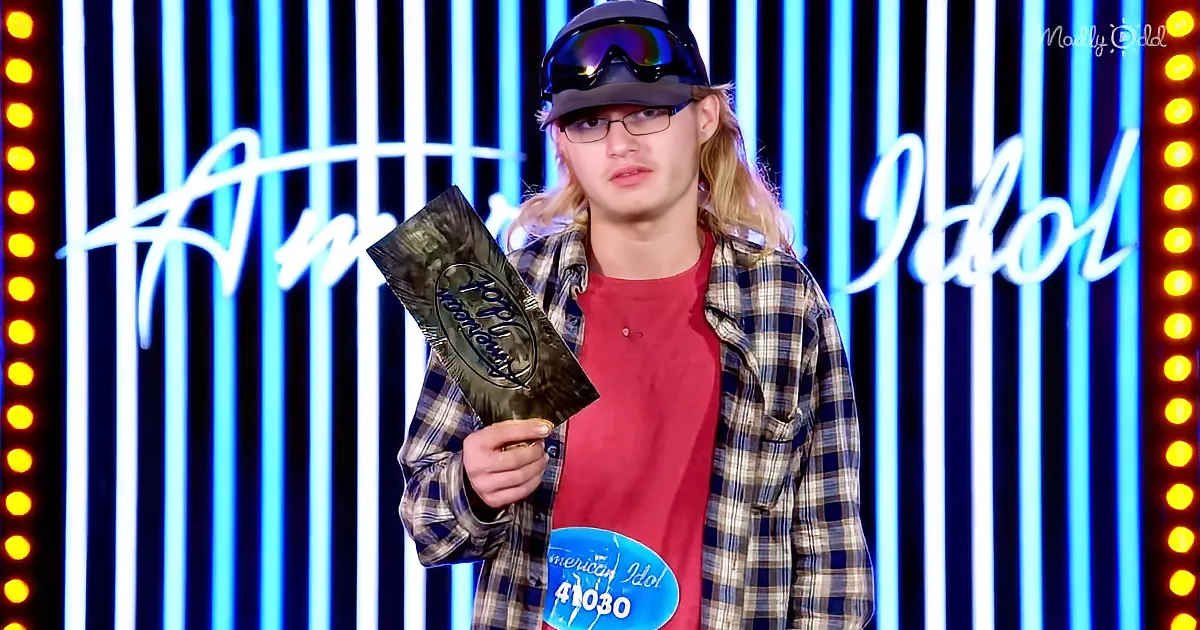 A person wearing a plaid shirt and goggles holds an "American Idol" sign, standing in front of a blue-lit backdrop.