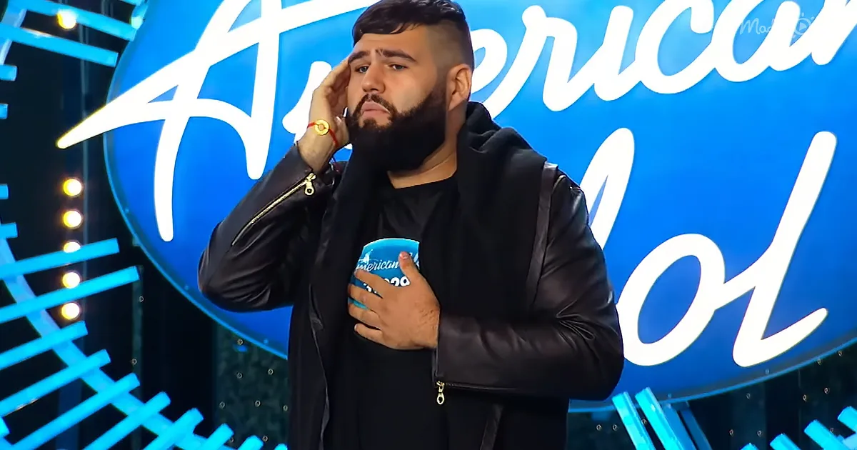 A man in a black jacket holds his hand to his head and chest, standing in front of an "American Idol" sign.