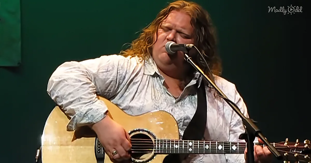 A person with long hair plays an acoustic guitar while singing into a microphone.