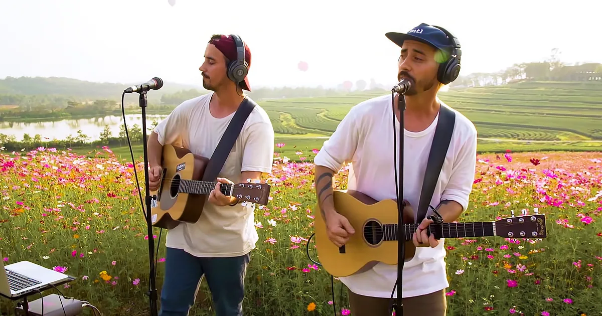 Two musicians play acoustic guitars in a field of flowers, surrounded by lush greenery and a serene lake.