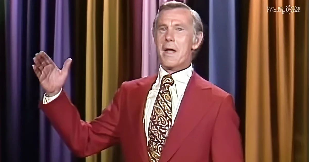 A man in a red suit and patterned tie gestures with his right hand against a colorful curtain backdrop.