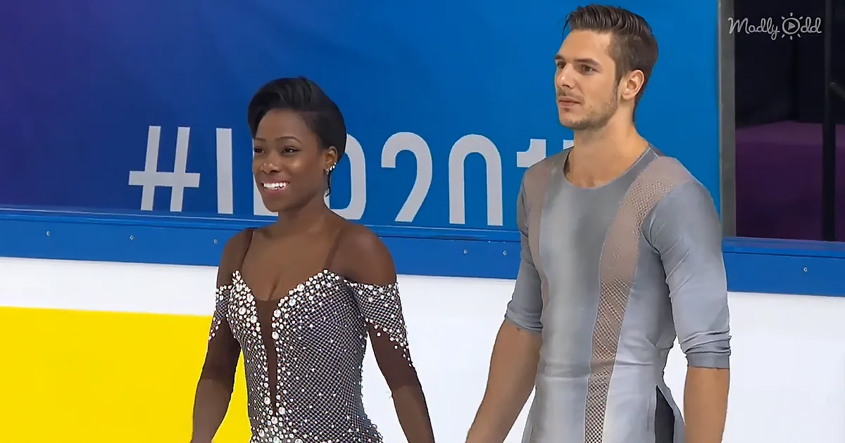 A figure skating pair stands on ice, woman in sparkly dress, man in mesh outfit, against a blue backdrop with "10201" and...