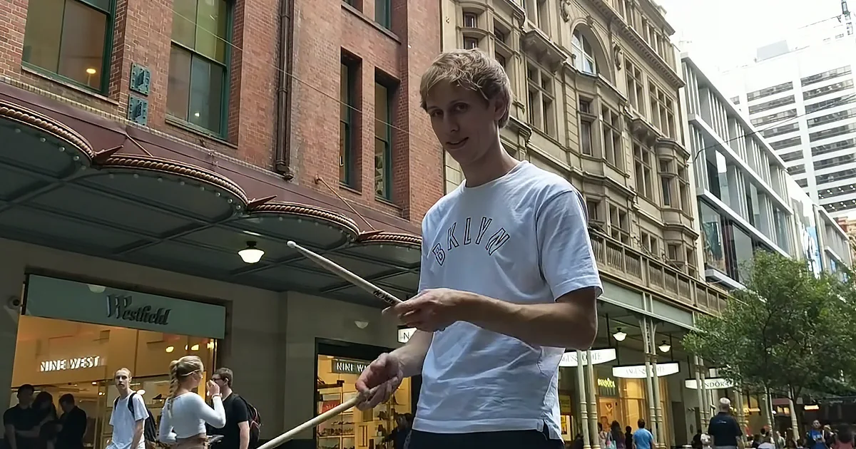 A person in a white "BKLYN" t-shirt holds drumsticks, standing on a city street with shops and pedestrians in the background.