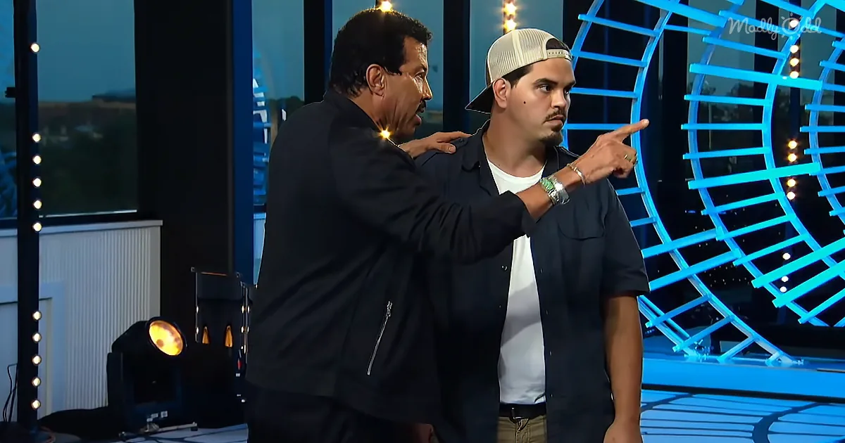 A man in a black jacket points to another man in a white shirt and cap on a stage with blue lighting.