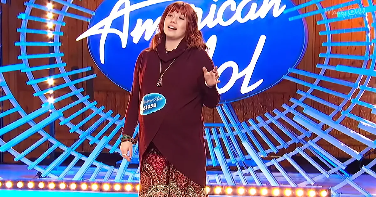 A person stands on stage in front of an "American Idol" backdrop, gesturing with their hands.