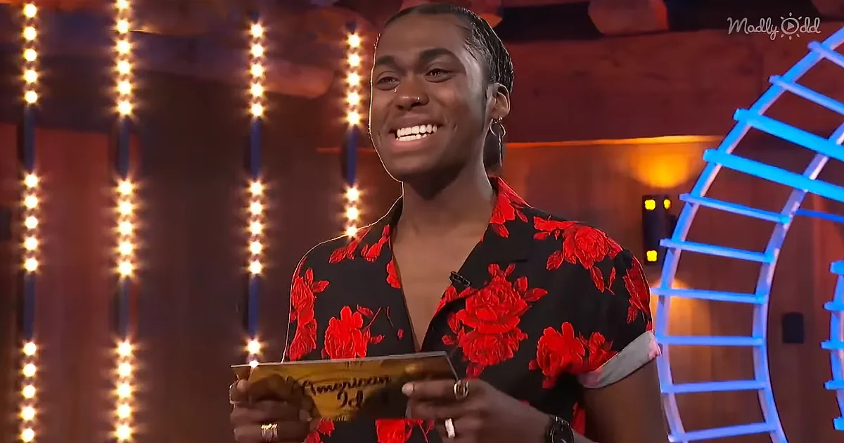 A person in a red floral shirt smiles while holding a card, standing in front of string lights and a blue-lit structure.
