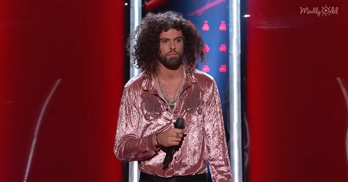 A man with curly hair and a beard, wearing a shiny pink shirt, holds a microphone on stage.