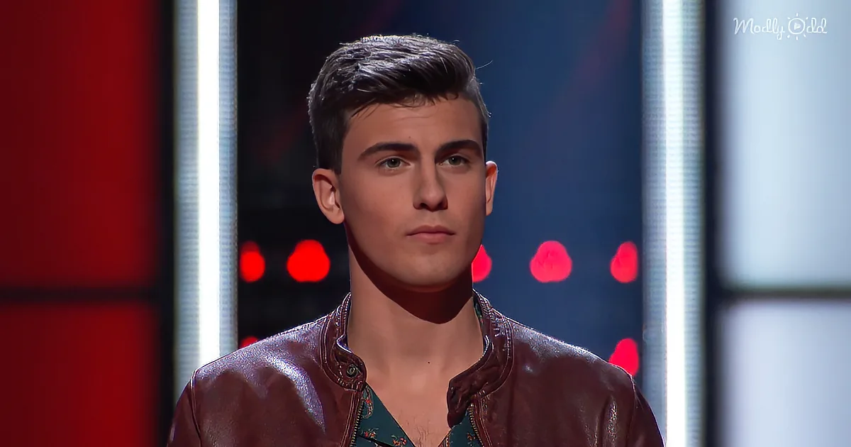 A person in a brown leather jacket stands against a red and white backdrop, looking forward.