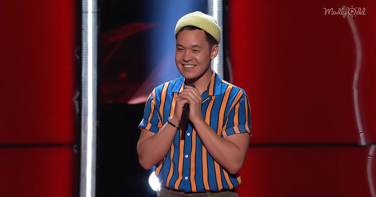 A man in a striped shirt and beanie smiles while holding a microphone on stage.