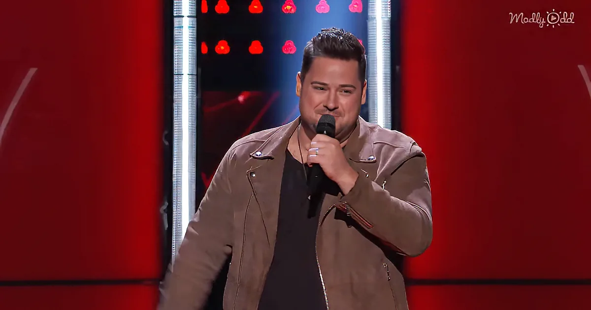 A man in a brown jacket holds a microphone on stage with red and white lights behind him.
