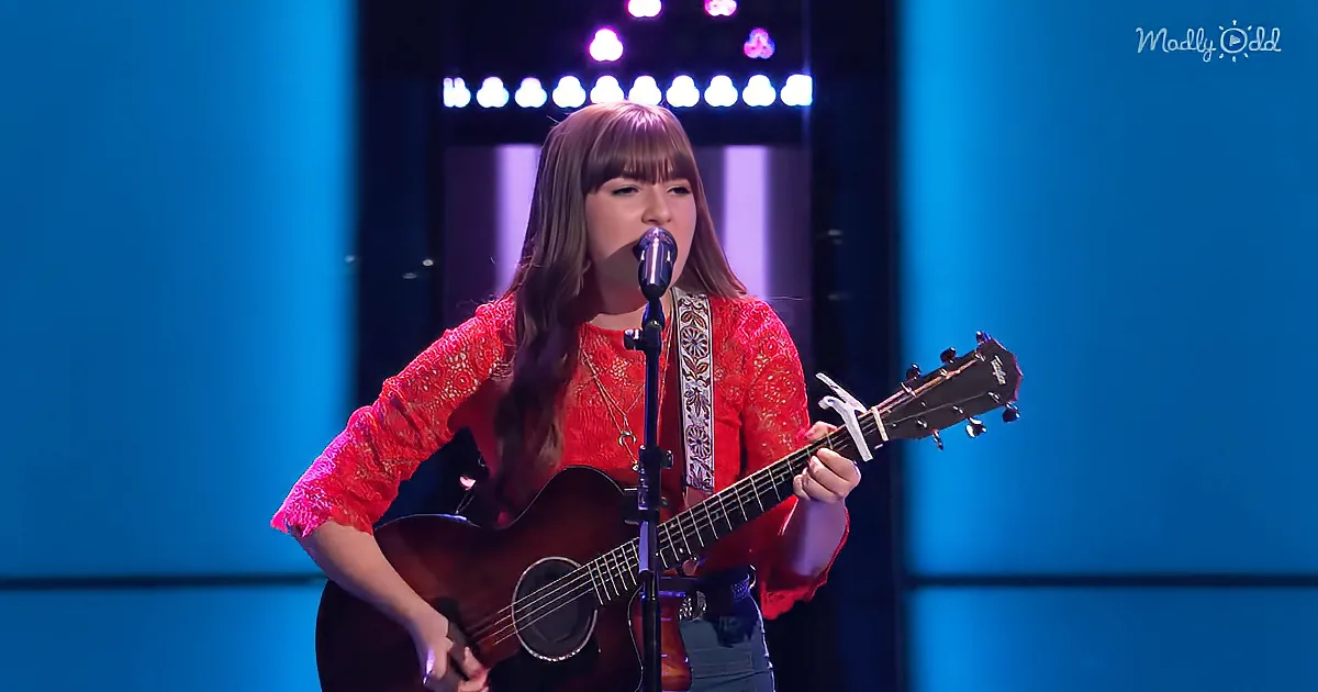 A woman in a red lace top plays an acoustic guitar and sings into a microphone on stage.