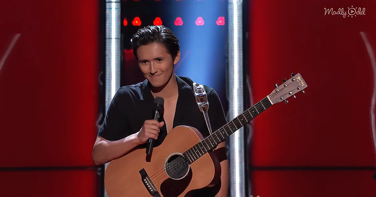 A man in a black shirt holds a microphone and an acoustic guitar on stage with red and blue lighting.