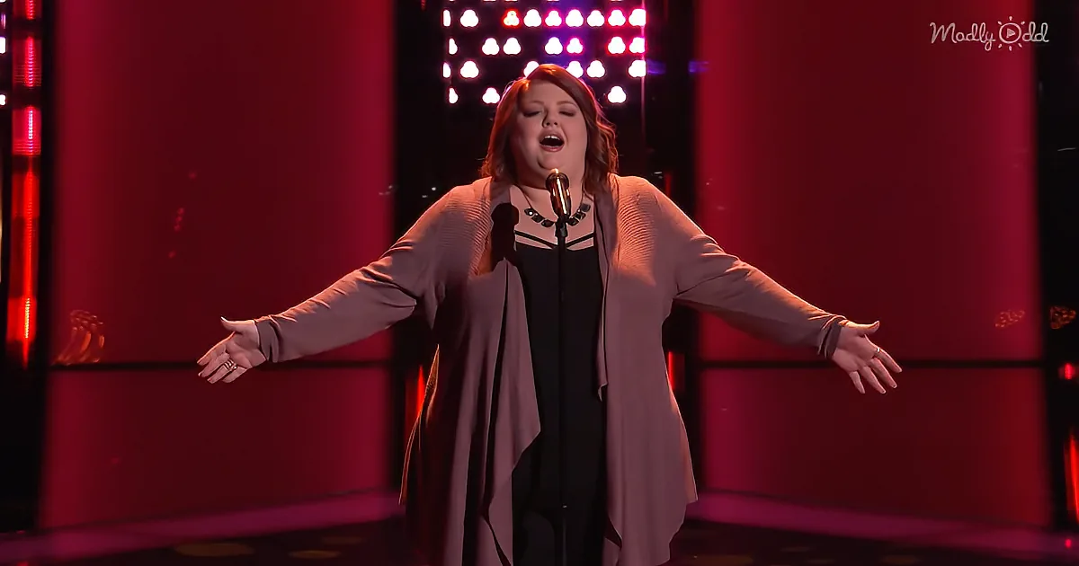 A woman stands on stage with arms outstretched, singing into a microphone, wearing a beige cardigan and black dress.