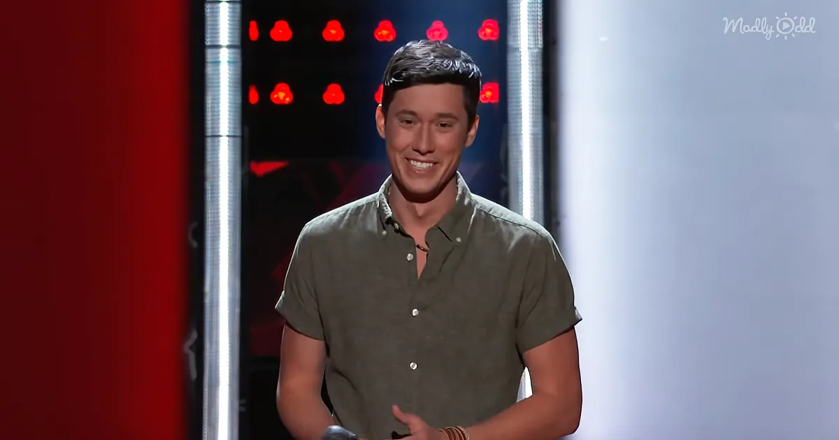 A man in a green shirt smiles on stage with red lights and a spotlight behind him.