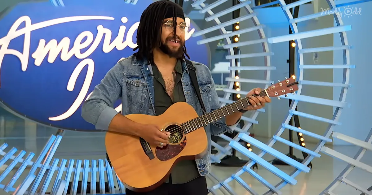 A man with dreadlocks and glasses plays an acoustic guitar in front of an "American Idol" sign.