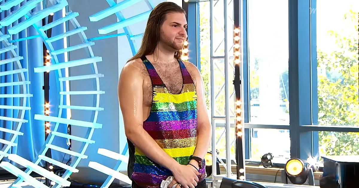 A man in a rainbow sequined tank top stands in front of a blue light structure, looking to the side.