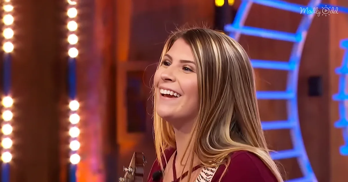 A smiling woman with long blonde hair, wearing a maroon top, sits in front of a backdrop with vertical lights and a blue...