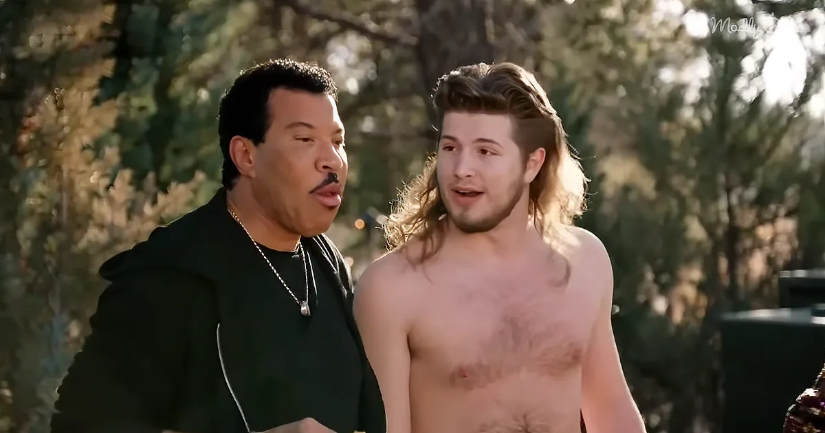 A shirtless man with long hair and a mustache stands next to a man in a black hoodie, both outdoors with trees in the...