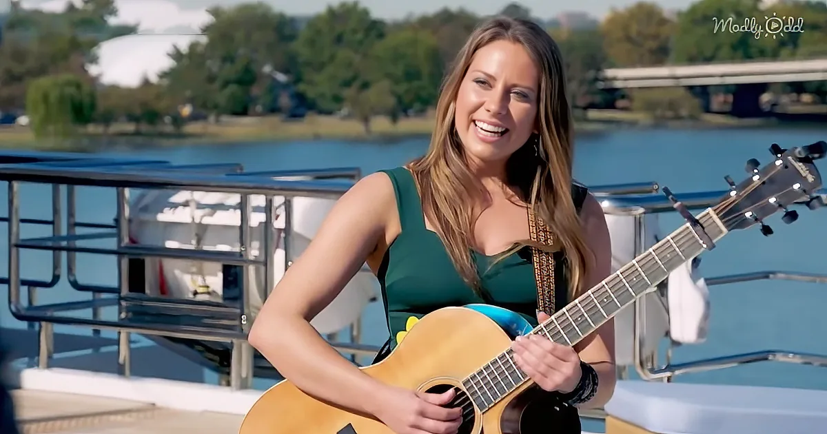A woman in a green dress smiles while playing an acoustic guitar on a boat with a scenic background.