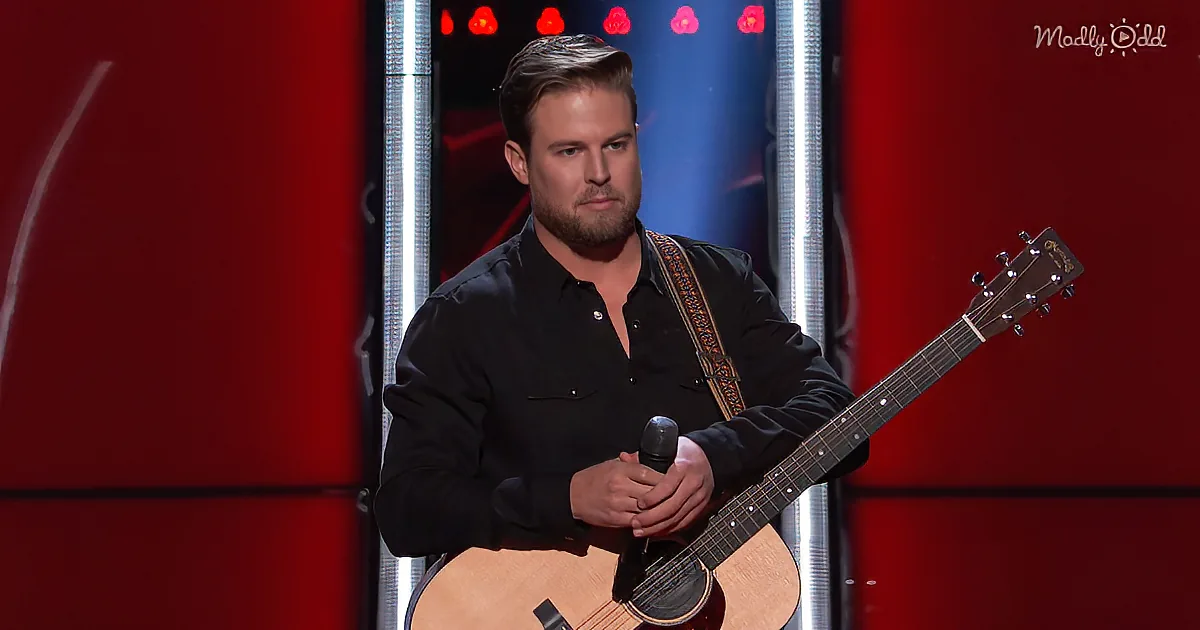 A man in a black shirt holds a guitar and microphone on stage with red lights behind him.