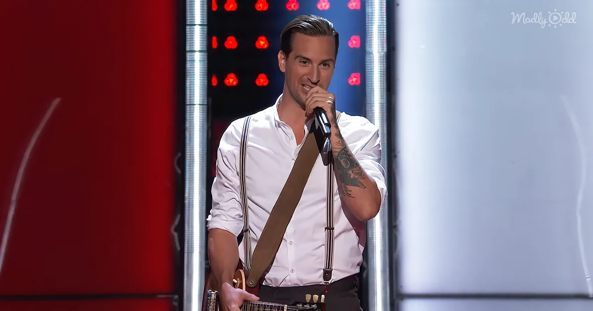 A man in a white shirt holds a microphone and guitar, standing on a stage with red and white lights.