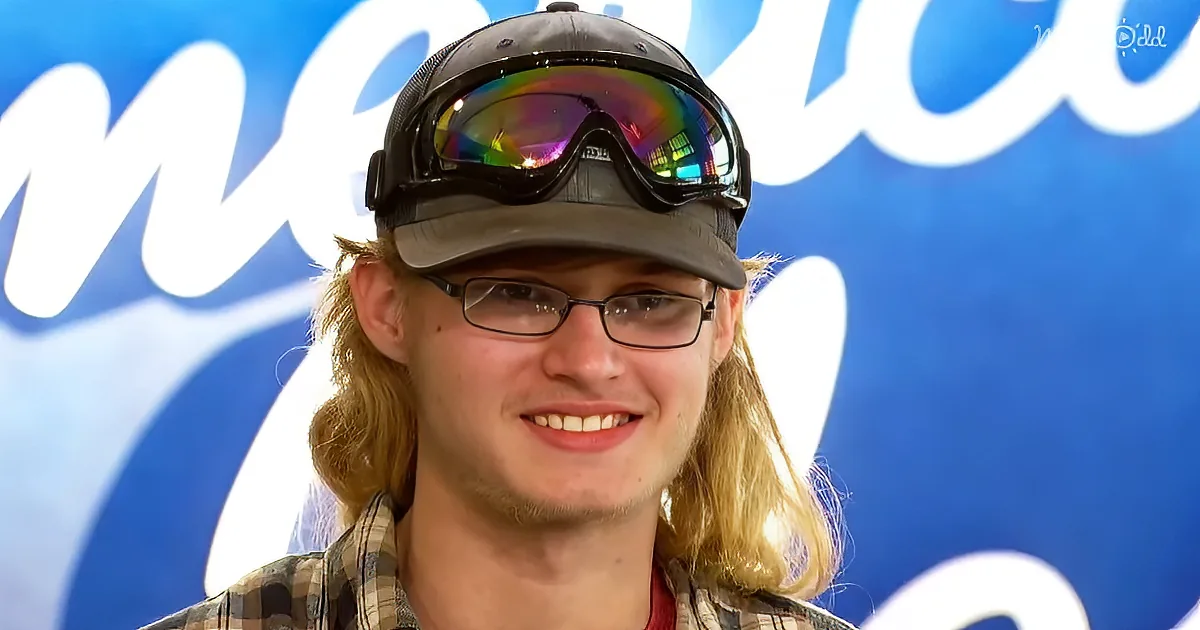 A smiling person with long hair, glasses, a cap, and goggles, wearing a plaid shirt, against a blue background.