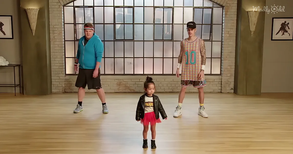 A young girl in a red skirt and black jacket stands between two men in a gym setting.