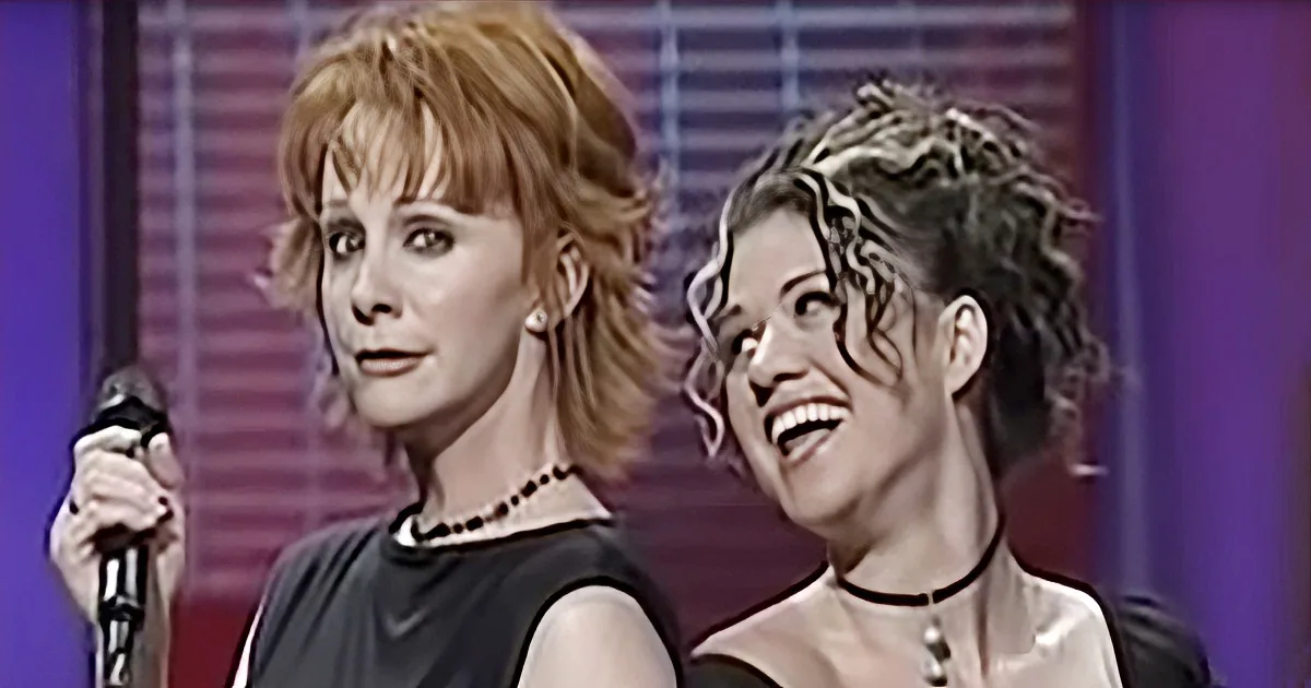 Two women, one holding a microphone, both wearing black dresses and necklaces, smiling and looking to the side.