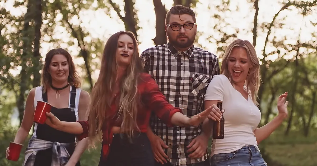 A woman in a red shirt dances with friends in a park, holding a beer bottle.