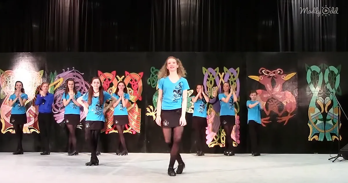 A young woman in a blue shirt and black skirt dances on stage, surrounded by a group of women in matching outfits.