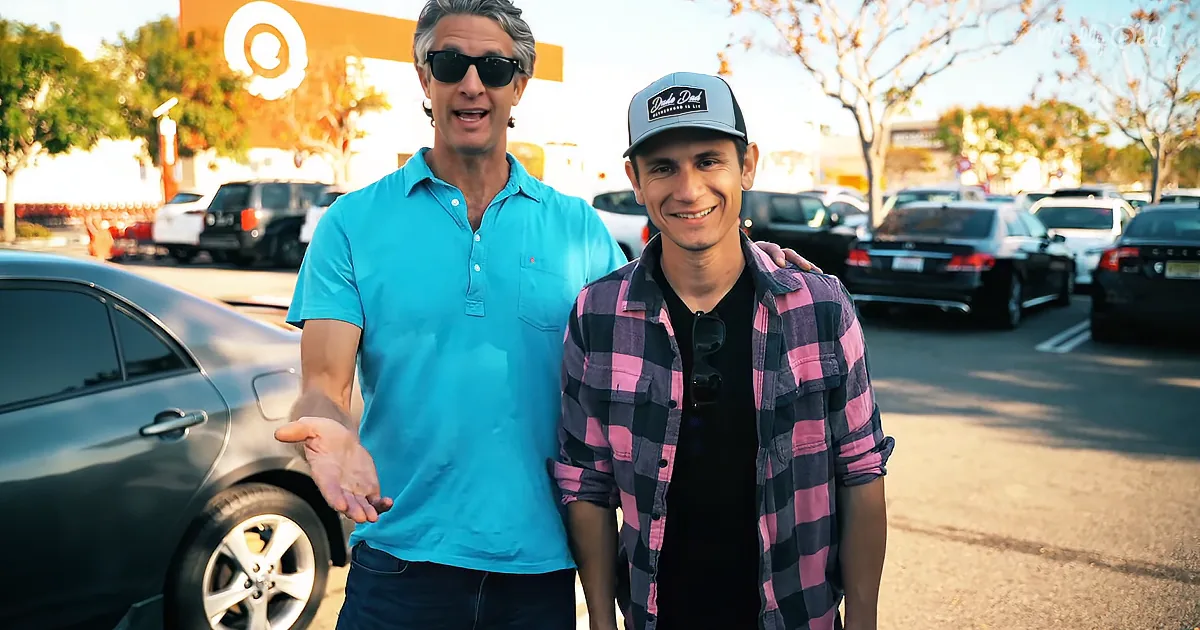 Two men stand in a parking lot, one in a blue shirt gesturing, the other in a plaid shirt smiling.