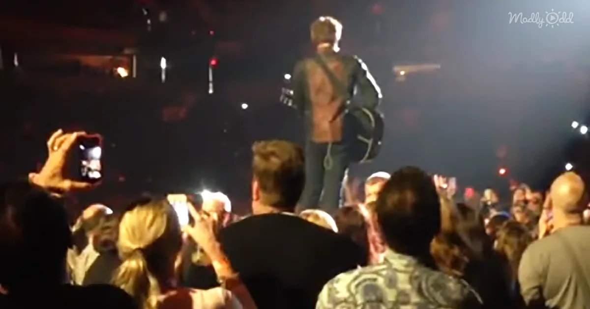 A person stands on stage holding a guitar, surrounded by a crowd taking photos and videos.
