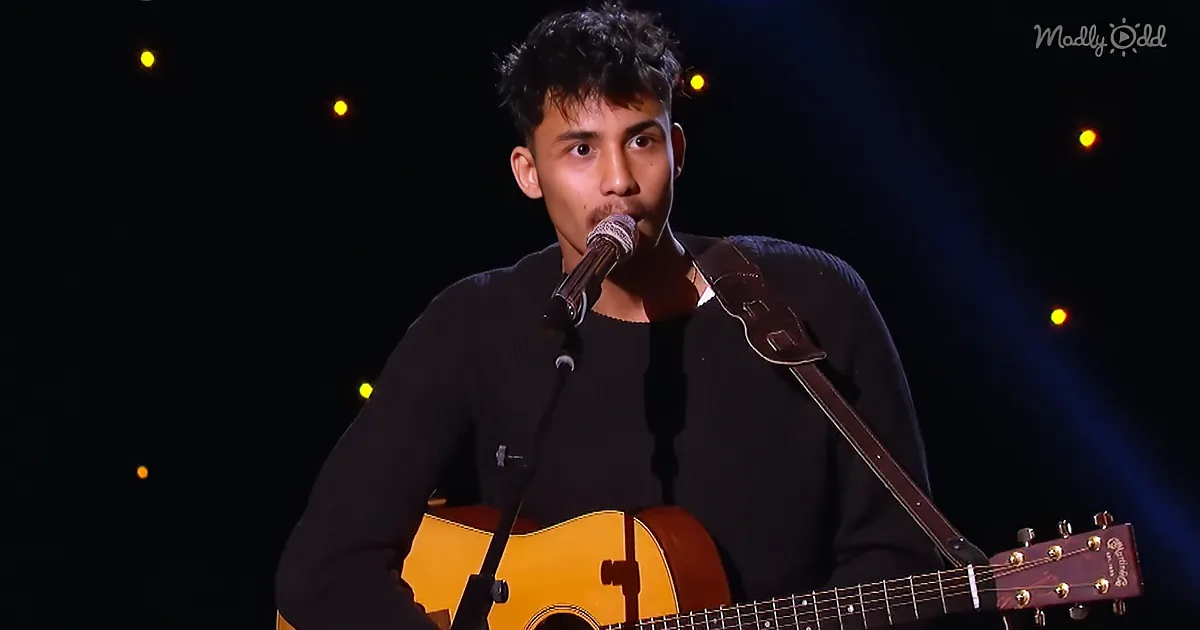 A man sings into a microphone while holding an acoustic guitar on stage.