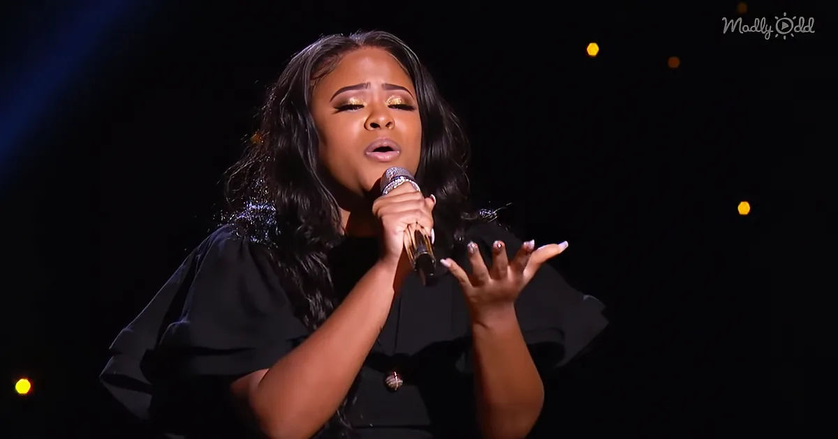 A woman in black attire sings into a microphone, her hand raised, with a dark background and yellow lights.