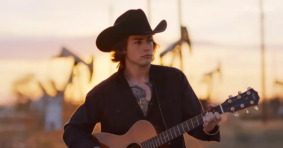 A person in a cowboy hat plays an acoustic guitar against a sunset backdrop with oil pumps.