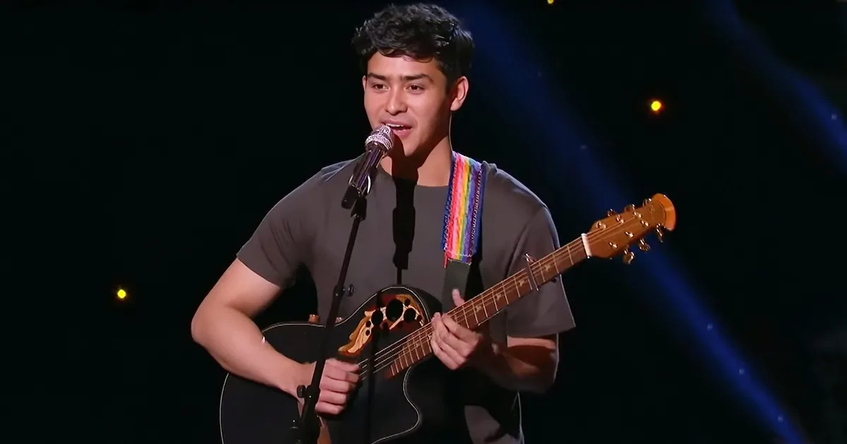 A person plays guitar and sings into a microphone on stage with a colorful scarf.