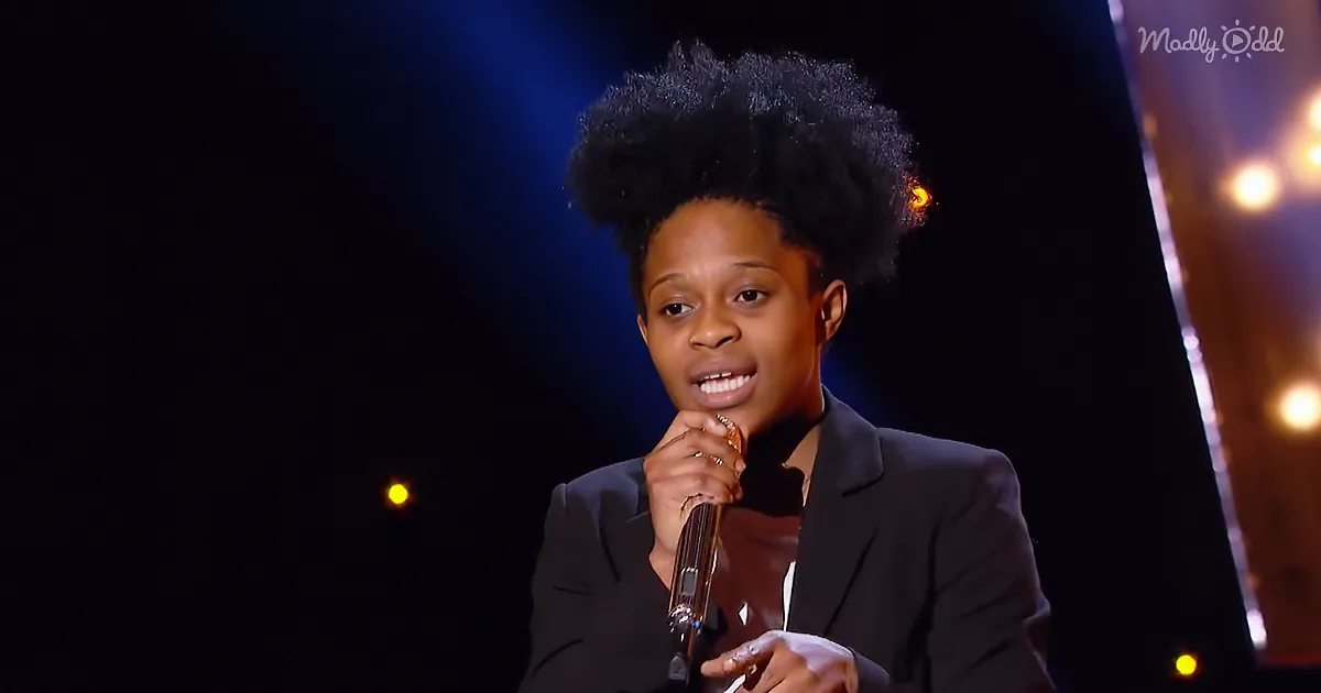 A person with a large afro hairstyle holds a microphone on stage, wearing a dark suit, with stage lights in the background.