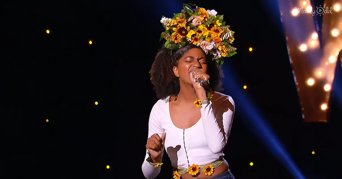 A woman sings passionately on stage, adorned with a floral crown and matching accessories, illuminated by stage lights.