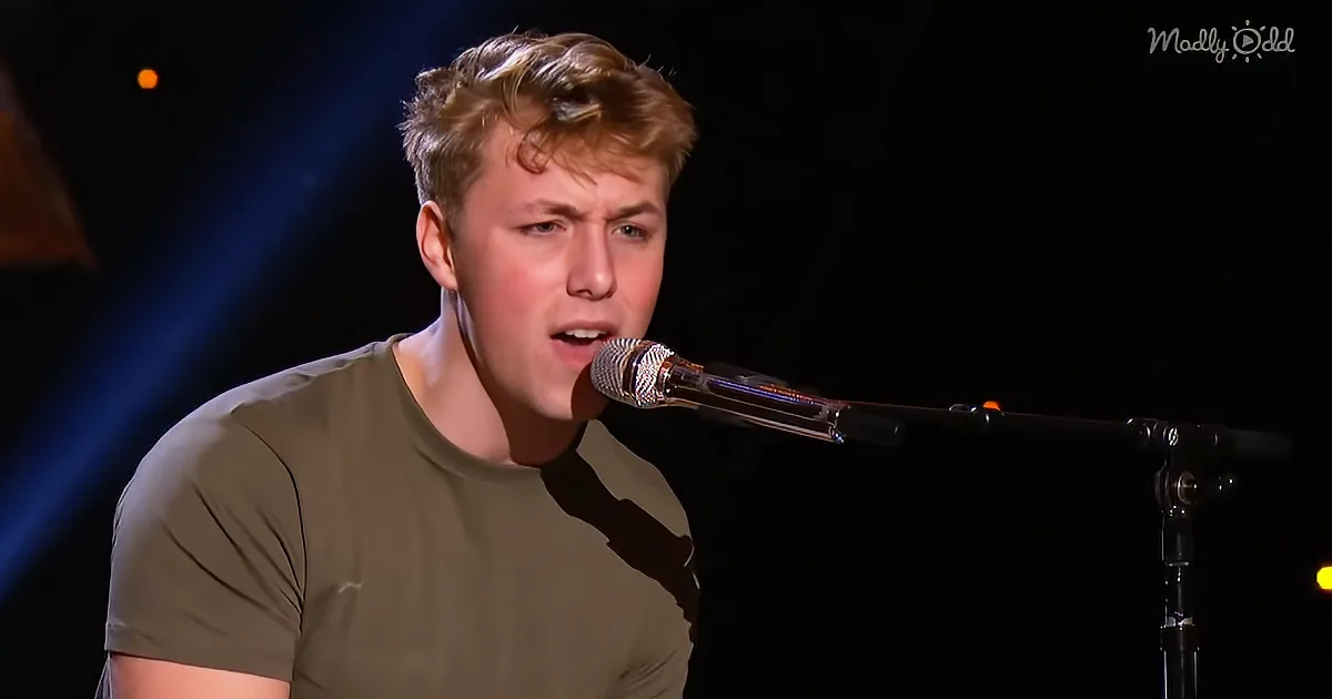 A person in a green shirt sings into a microphone on stage with a dark background and blue light.