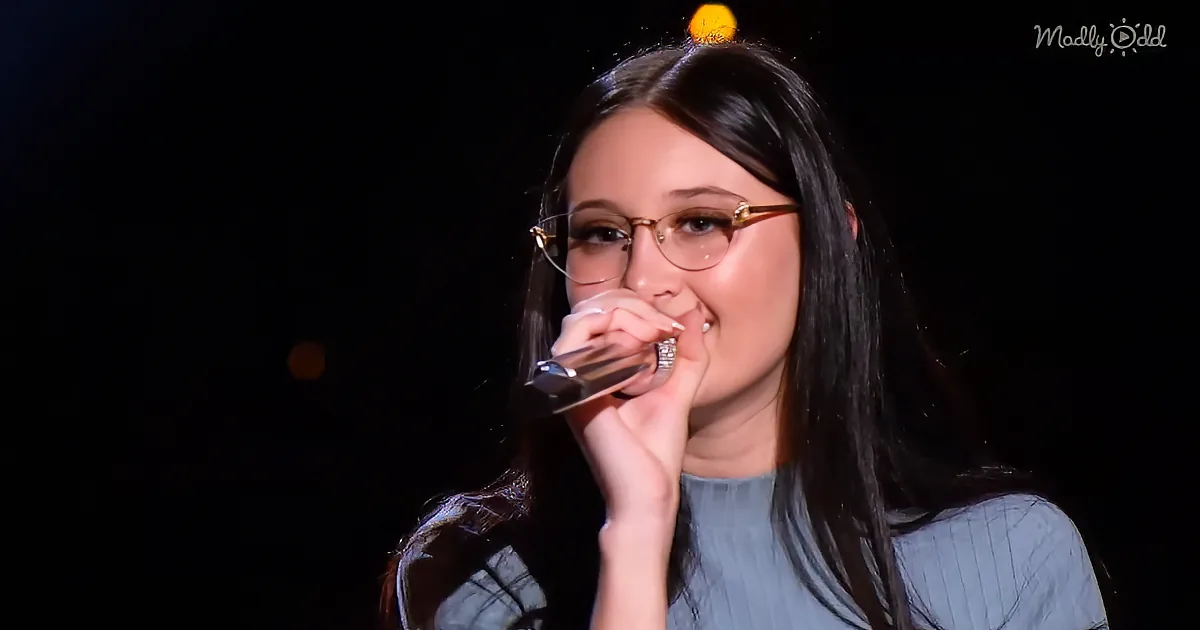 A woman with long dark hair and glasses holds a microphone close to her mouth, singing.