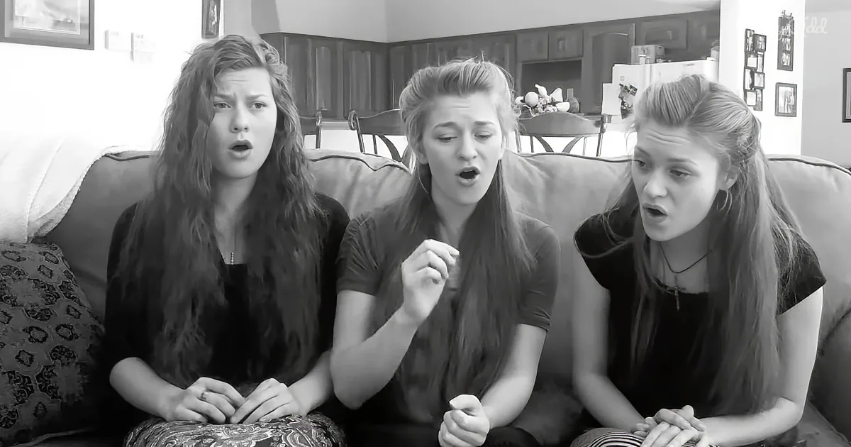 Three young women sit on a couch, appearing surprised or shocked, in a black-and-white image.