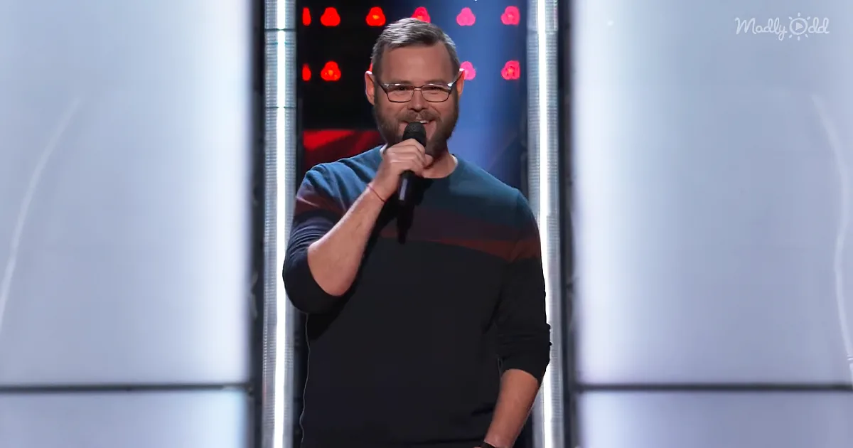 A man with glasses holds a microphone on stage, wearing a striped sweater, with red lights and white panels in the...