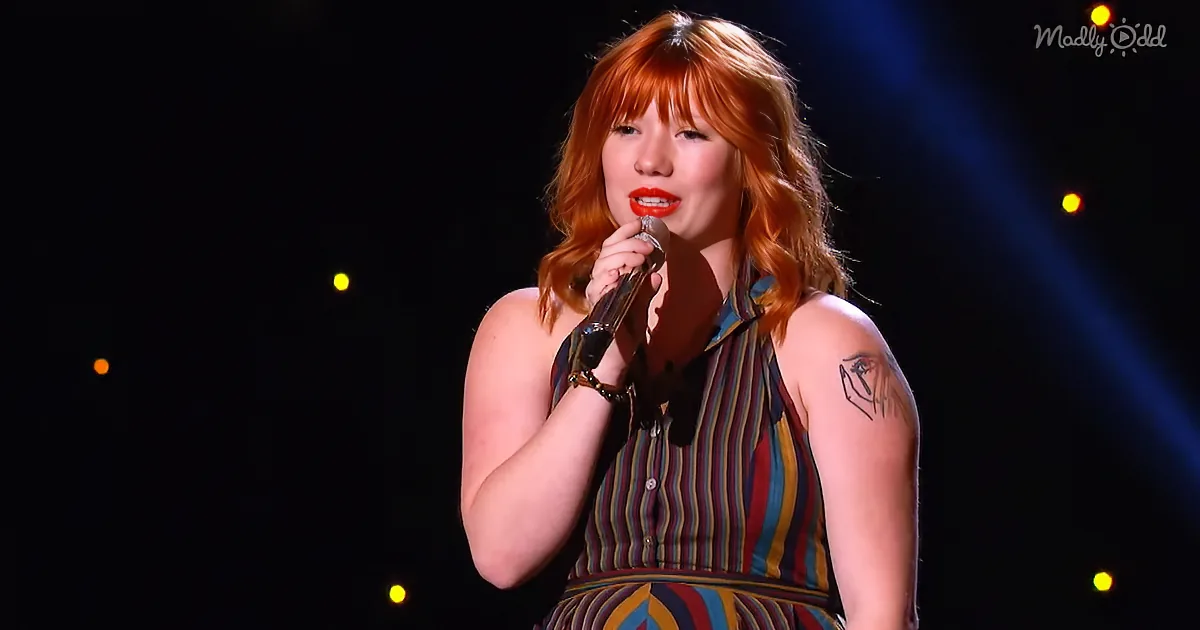 A woman with red hair and a striped dress sings into a microphone on stage.