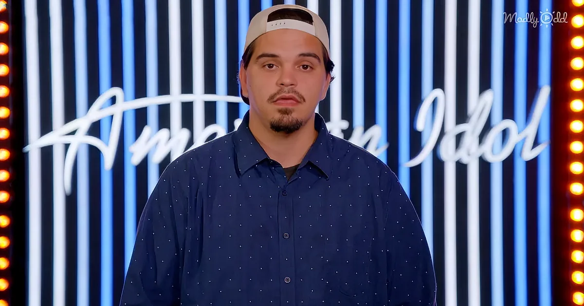 A man in a blue polka-dot shirt and cap stands in front of a striped "American Idol" backdrop.