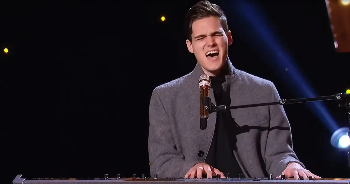 A person in a gray suit sings into a microphone on stage, with a keyboard in front.