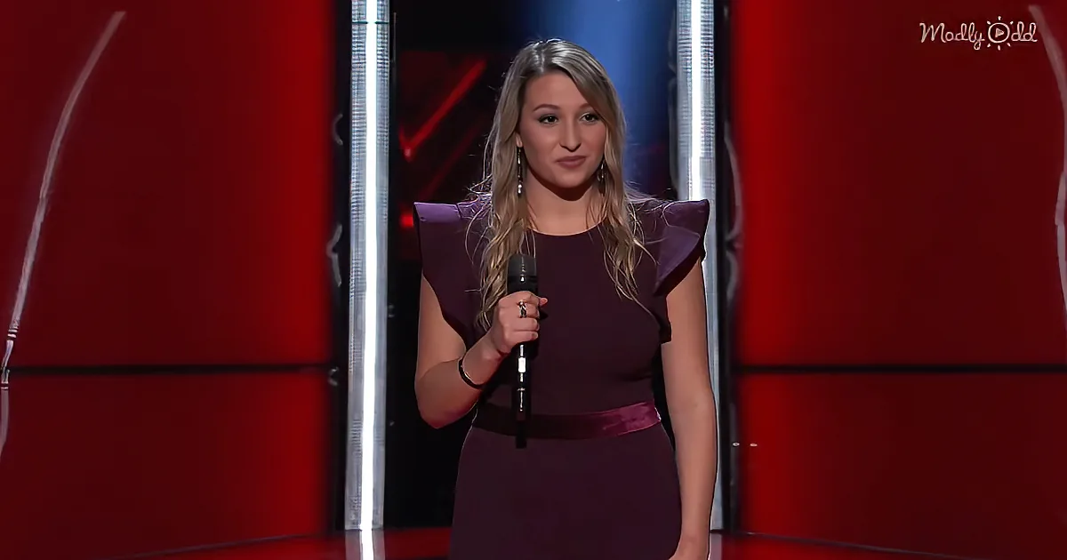 A woman in a purple dress holds a microphone on a stage with red and black lighting.