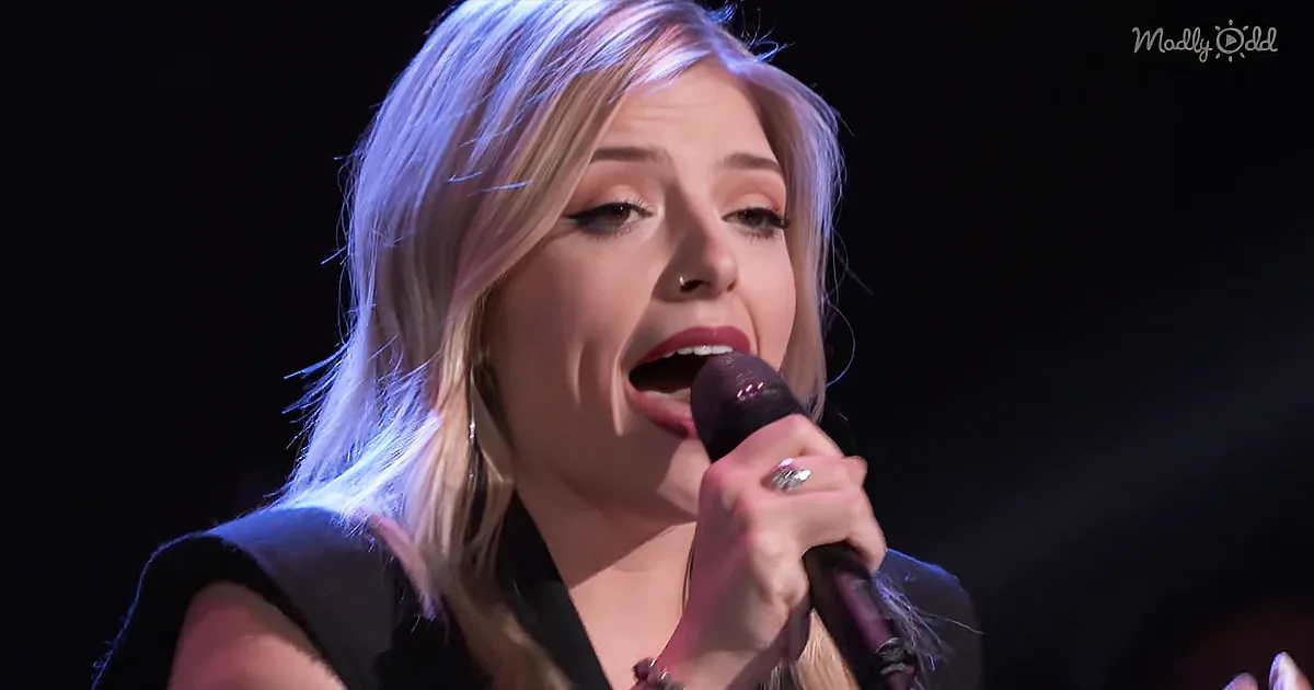 A woman with blonde hair sings into a microphone, wearing a black top and a ring, against a dark background.