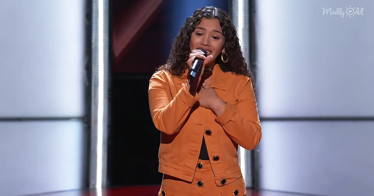 A woman in an orange outfit sings into a microphone on a stage with a spotlight.