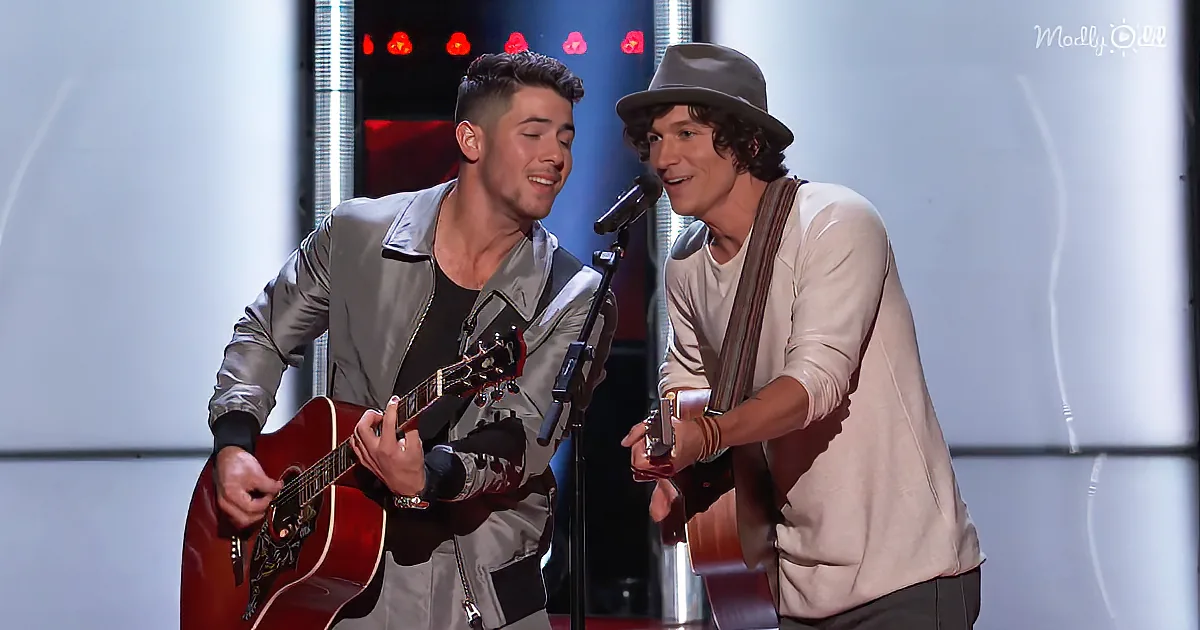 Two men on stage, one playing guitar, both smiling, one wearing a hat.